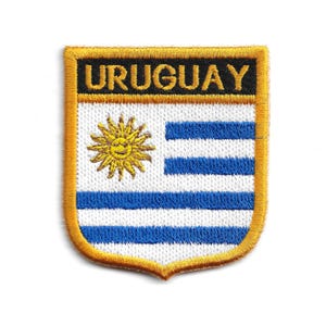 Pode incluir: Patch bordado com a bandeira do Uruguai. O patch tem uma borda dourada e formato de escudo. O design da bandeira inclui listras azuis e brancas, um sol amarelo e a palavra "URUGUAY" em preto.