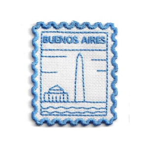 Puede incluir: Parche bordado con un diseño azul y blanco de un monumento de Buenos Aires, que se asemeja a un sello postal. El texto "BUENOS AIRES" está en la parte superior. El parche tiene un borde azul festoneado.