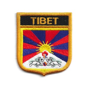 Puede incluir: Parche bordado con la palabra "TIBET" en letras doradas sobre fondo negro. El parche en forma de escudo muestra un sol, rayos rojos y azules y dos leones de nieve. El borde es dorado.