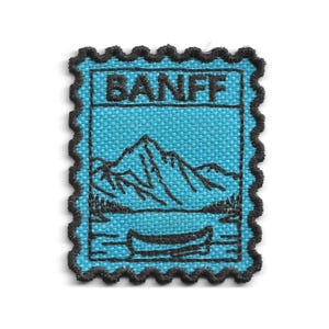 Può includere: Una toppa ricamata rettangolare con bordo nero smerlato su sfondo azzurro. Il disegno raffigura una catena montuosa sopra un lago con una canoa, e la parola BANFF è ricamata in nero nella parte superiore della toppa.