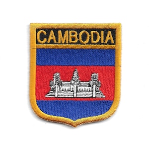 Może przedstawiać: Haftowana naszywka z napisem "CAMBODIA" w kolorze złotym nad niebiesko-czerwonym wzorem w paski. Naszywka zawiera biało-szarą ilustrację świątyni. Ma złotą obwódkę i kształt tarczy.