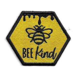 Może przedstawiać: Żółty sześciokątny łata z czarnym obramowaniem. Łata przedstawia czarną pszczołę z żółtymi paskami i tekst "BEE KIND" w kolorze czarnym.
