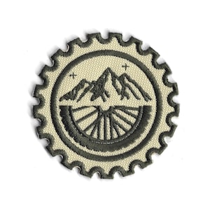Könnte beinhalten: Runder, gestickter Aufnäher mit einem Berg- und Fahrradfelgen-Design. Der Aufnäher hat einen cremefarbenen Hintergrund und einen dunkelgrauen Rand. Die Bergkette ist in der Mitte dargestellt, über einem Fahrradfelgen.