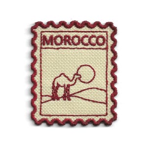 May include: A rectangular embroidered patch with a scalloped edge in dark red thread on a cream-coloured fabric base. The design features the word MOROCCO in dark red capital letters above a line drawing of a camel standing in a desert landscape.
