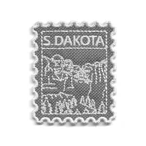 May include: A gray embroidered patch in the shape of a postage stamp. The patch features the text "S. DAKOTA" at the top and a detailed white embroidered design of a landscape. The edges are scalloped with white thread.