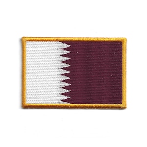 Puede incluir: Parche rectangular con la bandera de Qatar. El diseño presenta un campo granate a la derecha y una sección blanca a la izquierda, separados por un borde dentado. Un borde dorado enmarca el diseño.