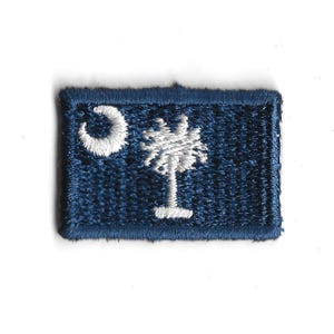 May include: A blue embroidered patch featuring the South Carolina state flag. The flag depicts a white crescent moon and palmetto tree on a blue background.