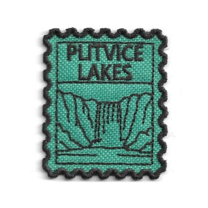 Puede incluir: Un parche bordado rectangular con borde festoneado, que presenta un fondo verde azulado y costuras negras. El diseño representa un paisaje de cascada con el texto PLITVICE LAKES bordado en hilo negro en la parte superior del parche.
