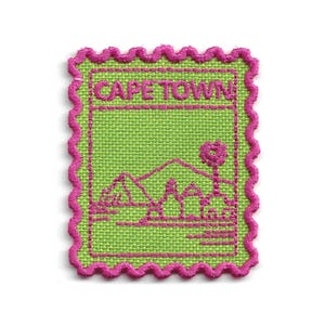 Peut inclure: Un écusson brodé rectangulaire avec un bord festonné rose vif, sur un fond vert lime. Le texte CAPE TOWN est brodé en rose en haut, au-dessus d'une illustration paysagère stylisée représentant des montagnes et un palmier en rose.