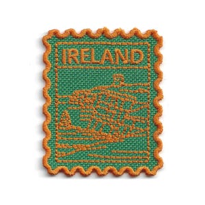 May include: A rectangular embroidered patch with a scalloped orange border and a green fabric background. The word IRELAND is stitched in orange at the top, above an orange embroidered graphic depicting a traditional Irish cottage landscape.