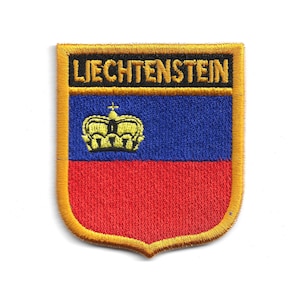 Puede incluir: Parche bordado con la bandera de Liechtenstein. El parche tiene forma de escudo con un borde dorado. La mitad superior es azul, la mitad inferior es roja y una corona dorada está en la sección azul. La palabra "LIECHTENSTEIN" está bordada en dorado.