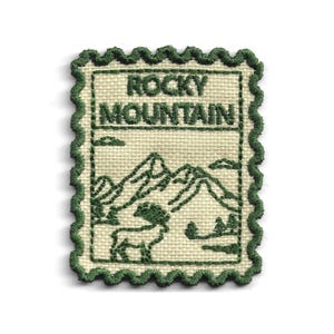 May include: A rectangular embroidered patch with a scalloped edge, featuring a cream-colored fabric base and dark green thread. The design depicts a mountain range, a bighorn sheep, and the text "ROCKY MOUNTAIN" in a bold, block font.