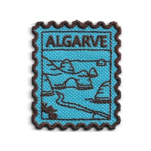 May include: A rectangular embroidered patch with a scalloped brown border on a light blue background. The design features a dark brown stitched outline of a coastal rock formation and the word ALGARVE in dark brown capital letters at the top.