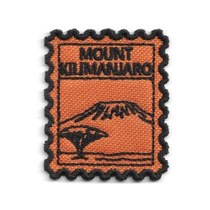 Puede incluir: Un parche bordado rectangular con un borde negro festoneado. El parche presenta un fondo de tela naranja con costuras negras que representan el monte Kilimanjaro y un árbol, con el texto MOUNT KILIMANJARO bordado en color negro.