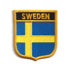 Op de afbeelding: Een schildvormige patch met de Zweedse vlag. Het ontwerp bevat een gele kruis op een blauwe achtergrond, met het woord "SWEDEN" in gele letters op een zwarte banner. De patch heeft een gouden rand.