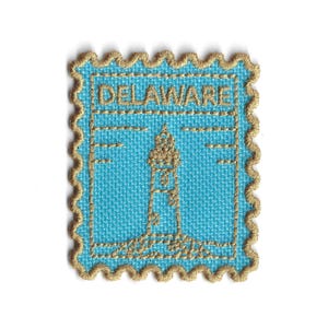 以下が含まれることがあります： 水色の背景に、金色の灯台のデザインと「DELAWARE」の文字が刺繍されたパッチ。 パッチには、切手のような装飾的な金の縁取りがあります。