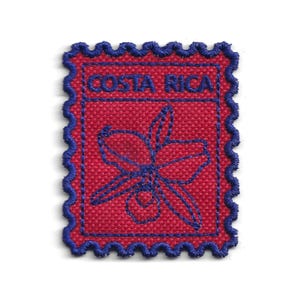 May include: A red fabric patch with a blue embroidered design of a flower and the words "COSTA RICA". The patch has a blue scalloped border, resembling a postage stamp. The design is centered on a white background.