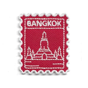Puede incluir: Parche rojo bordado con forma de sello postal. El parche presenta la palabra "BANGKOK" en letras blancas en la parte superior y un dibujo lineal blanco de un templo. Los bordes están festoneados con hilo blanco.