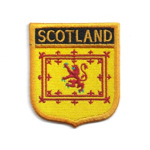 May include: Embroidered patch featuring the word "SCOTLAND" in black on a gold background. The patch has a shield shape with a yellow border. Inside is a yellow field with a red lion rampant and a red border.