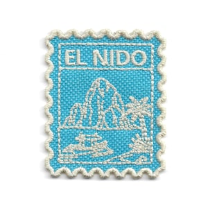 May include: A rectangular embroidered patch with a scalloped edge, featuring a bright blue background and white stitching. The design depicts a mountain landscape, a palm tree, and the text EL NIDO in a bold, sans-serif font at the top.