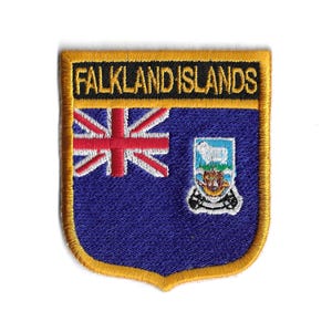 May include: An embroidered patch with a blue background and the text "FALKLAND ISLANDS" in black. The patch features the Falkland Islands flag with a Union Jack in the top left corner and the Falkland Islands coat of arms in the bottom right corner.