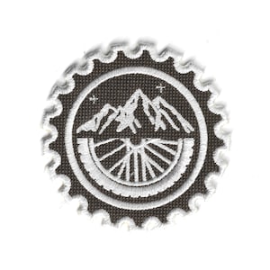 May include: Embroidered patch featuring a mountain range and a bicycle wheel design in white on a dark brown background. The patch has a circular shape with a white, scalloped border. Two small stars are visible.