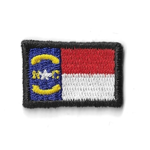May include: A small embroidered patch depicting the North Carolina state flag. The flag is blue with a yellow star and a yellow crescent moon in the centre. The flag is bordered by a red stripe and a white stripe.