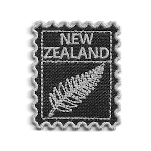 May include: A rectangular embroidered patch shaped like a postage stamp with a scalloped border. The patch features a black background with silver-grey stitching. It displays the text NEW ZEALAND above a silver-grey embroidered silver fern leaf.