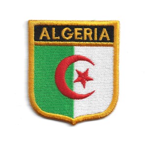 Op de afbeelding: Geborduurde patch met de Algerijnse vlag. De patch heeft een schildvorm met een gouden rand. Het vlagontwerp bevat een groene en witte verticale streep, een rode maan en ster, en het woord "ALGERIA" in het zwart.
