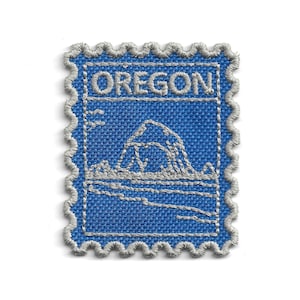 Puede incluir: Un parche de tela azul con un diseño bordado en gris de una montaña y la palabra "OREGON". El parche tiene un borde festoneado y recuerda a un sello postal.