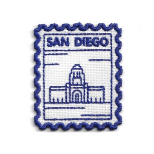 Puede incluir: Parche bordado con forma de sello postal, con el texto "SAN DIEGO" en la parte superior. El diseño incluye un contorno azul de un edificio. El parche tiene un fondo blanco y un borde azul festoneado.
