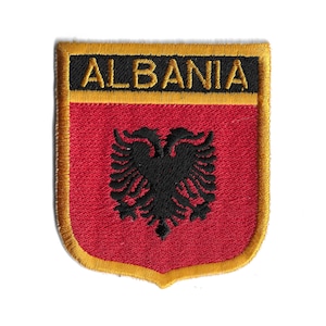 Puede incluir: Un parche bordado con fondo rojo y un águila bicéfala negra. El parche tiene un borde dorado y la palabra "ALBANIA" en letras negras.