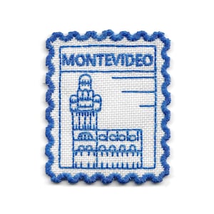 Pode incluir: Patch bordado com um design azul e branco com a palavra "MONTEVIDEO" no topo. Apresenta uma ilustração detalhada de um edifício ou monumento e uma borda ondulada azul. Provavelmente um item de lembrança ou com tema de viagem.