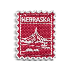 May include: A red embroidered patch in the shape of a postage stamp. The patch features the word "NEBRASKA" in white, along with a white illustration of a monument. The edges are scalloped with white thread.
