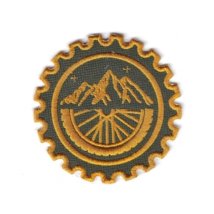 May include: Embroidered patch featuring a mountain range and bicycle wheel design in gold and olive green. The patch has a gear-shaped border. Ideal for outdoor enthusiasts and cyclists.