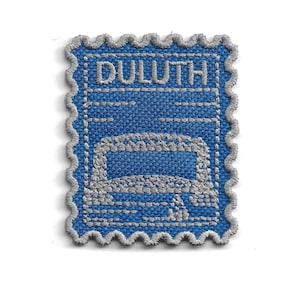 Peut inclure: Un écusson brodé rectangulaire en forme de timbre-poste avec une bordure argentée festonnée. Le tissu bleu présente un motif argenté brodé représentant un pont au-dessus de l'eau avec le mot DULUTH brodé en argent en haut au centre.
