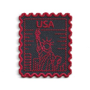 Könnte beinhalten: Ein rechteckiger, gestickter Aufnäher in Form einer Briefmarke mit dunkelgrauem Hintergrund und rotem Rand. Das Design zeigt die rote Stickerei der Freiheitsstatue und den Schriftzug USA in roten Großbuchstaben.