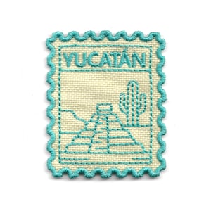 Puede incluir: Un parche de tela cuadrado, blanquecino, con un borde turquesa, que se asemeja a un sello postal. El parche presenta la palabra "YUCATAN" en turquesa, junto con una ilustración turquesa de una pirámide y un cactus.