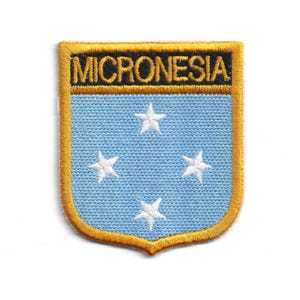 May include: Embroidered patch featuring the word "MICRONESIA" in gold lettering above a light blue shield with four white stars. The patch has a gold border and a shield shape.