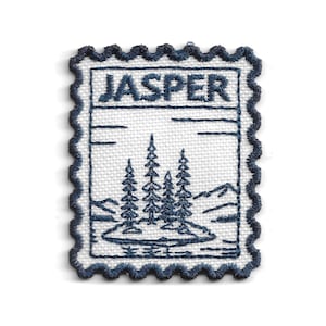 May include: A rectangular embroidered patch with a scalloped edge, featuring a white background and dark blue stitching. The design depicts a landscape with three pine trees, mountains, and the word JASPER embroidered in dark blue text.