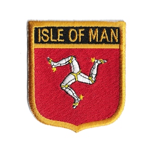May include: An embroidered patch featuring the Isle of Man flag. The flag is red with a yellow three-legged symbol, known as the triskelion, on a white background. The patch is surrounded by a gold border and has the words "Isle of Man" in black lettering.