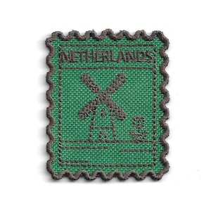 Puede incluir: Un parche bordado rectangular con un borde festoneado de color marrón oscuro sobre un fondo de tela verde. El diseño presenta un molino de viento y una pequeña flor bordados en marrón oscuro, con el texto NETHERLANDS en la parte superior.