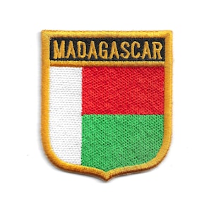 Könnte beinhalten: Gestickter Aufnäher mit der Flagge von Madagaskar. Der schildförmige Aufnäher hat einen goldenen Rand und das Wort "MADAGASCAR" oben. Das Flaggen-Design zeigt weiße, rote und grüne vertikale Streifen.