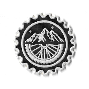 Op de afbeelding: Een cirkelvormige zwart-witte geborduurde patch met een bergketen en een fietswielontwerp. De patch heeft een geschulpte rand en twee kleine sterren. Geschikt voor kleding of accessoires.