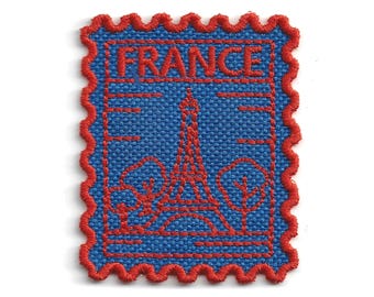 Puede incluir: Un parche bordado rectangular con forma de sello postal y borde rojo festoneado. El diseño presenta un fondo de tela azul con costuras de hilo rojo que representan la Torre Eiffel, dos árboles y la palabra FRANCE en la parte superior.
