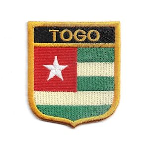 Op de afbeelding: Geborduurde patch met de vlag van Togo. De patch heeft de vorm van een schild met een rood vierkant en een witte ster, groene en witte strepen en het woord "TOGO" in gouden letters op een zwarte achtergrond. De patch is omrand met gouddraad.