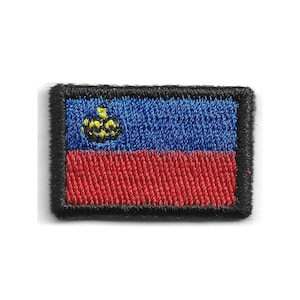 May include: A rectangular embroidered patch featuring the flag of Liechtenstein. The design consists of a horizontal blue band on top and a red band on the bottom, with a yellow crown emblem on the left side of the blue section, all bordered by black.