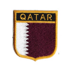 Könnte beinhalten: Gestickter Aufnäher mit dem Wort "QATAR" in Schwarz auf einem goldumrandeten schwarzen Rechteck. Der Aufnäher hat einen weinroten Hintergrund mit einem weißen, gezackten Randdesign auf der linken Seite und einen goldenen Rand.