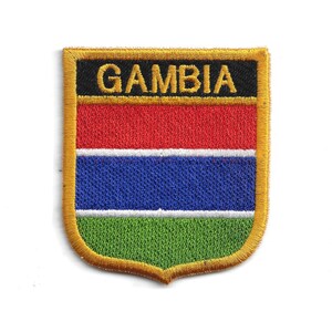 May include: Embroidered patch featuring the flag of Gambia. The patch has a shield shape with a gold border. The flag design includes horizontal stripes of red, blue, and green, with the word "GAMBIA" in gold letters on a black bar.