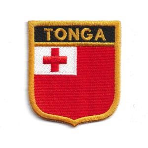 Op de afbeelding: Geborduurde patch met de vlag van Tonga. De patch heeft een rode achtergrond met een witte vierkant in de linkerbovenhoek, met een rood kruis. Het woord "TONGA" is in het wit geborduurd op een zwarte banner bovenaan, met een gouden rand.